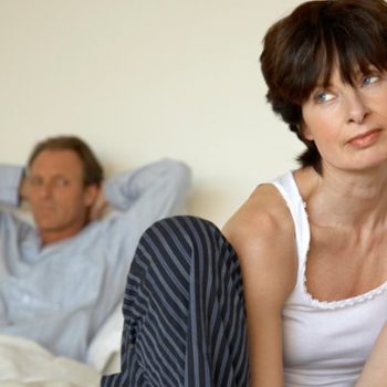 Woman looking uninterested in bed while man sits back relaxed in bed with his hands behind his head