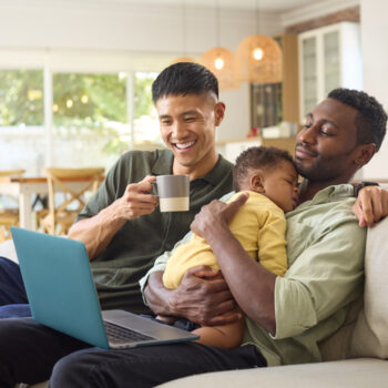 Same Sex Male Couple With Sleeping Baby Daughter On Sofa At Home Working On Laptop